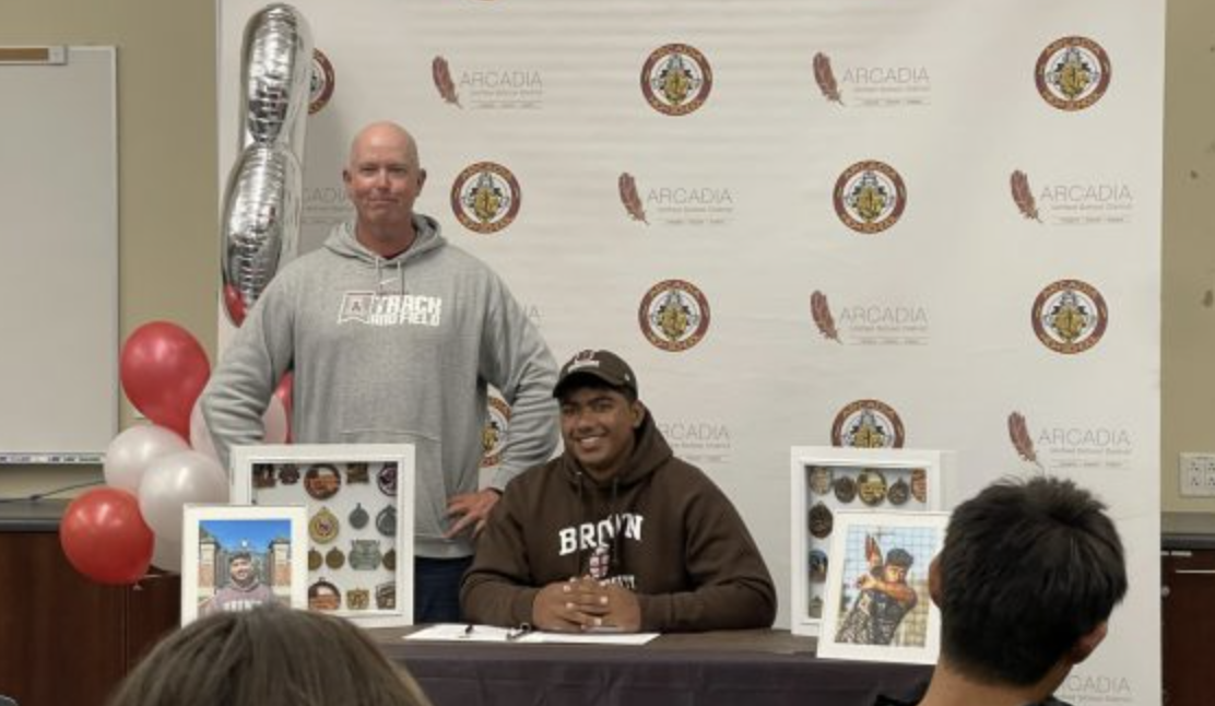 Track and Field Coach Christopher Schultz stands next to Biju as he signs. 